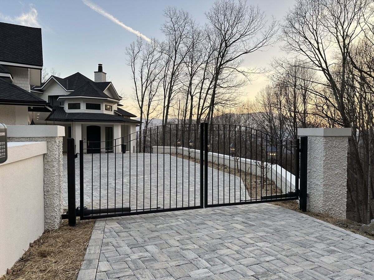 Black aluminum driveway gate hung between stone-faced columns at a paver drive entrance