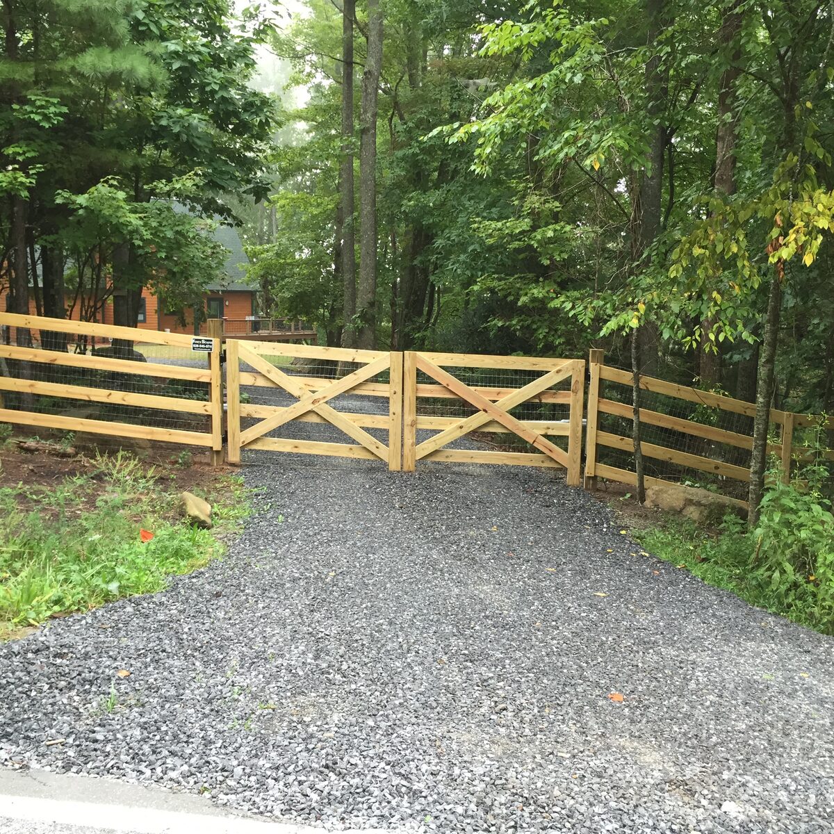 Cross-buck cedar double drive gate backed with wire mesh on a gravel driveway, matching four-rail fence running into the woods on both sides