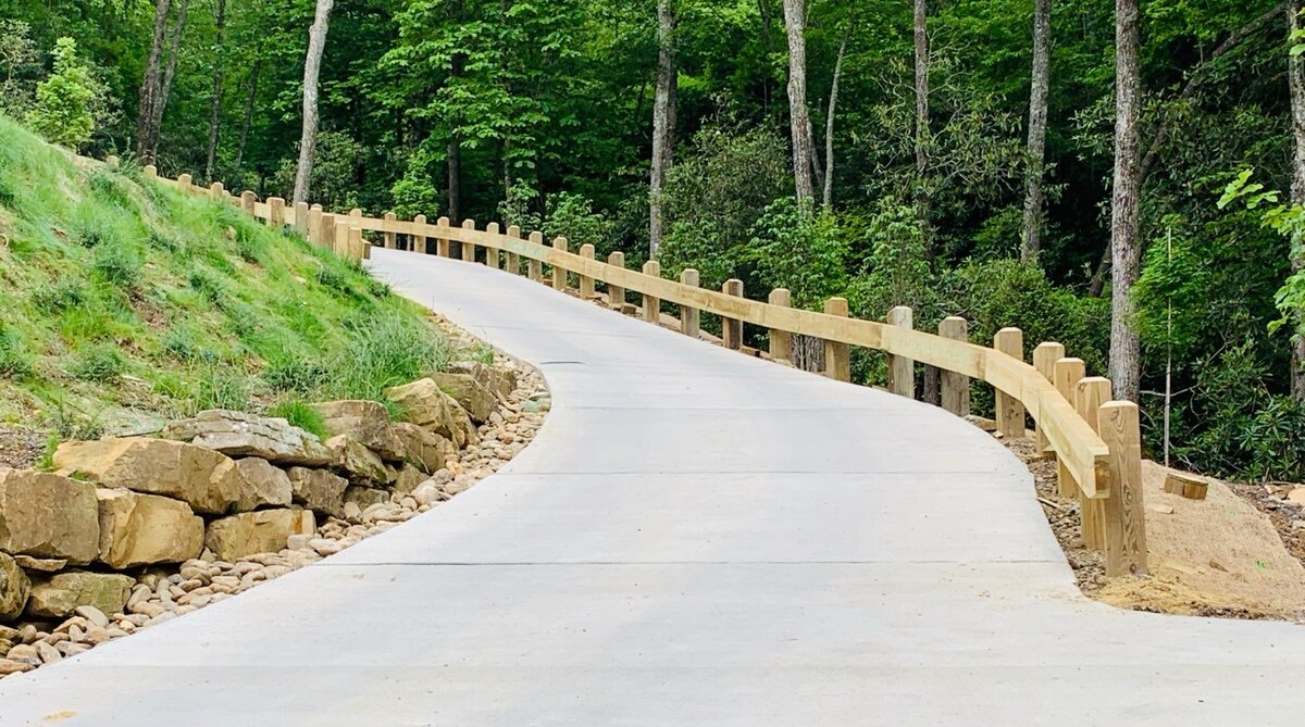 Timber post-and-rail guardrail along a curved concrete driveway on a wooded hillside